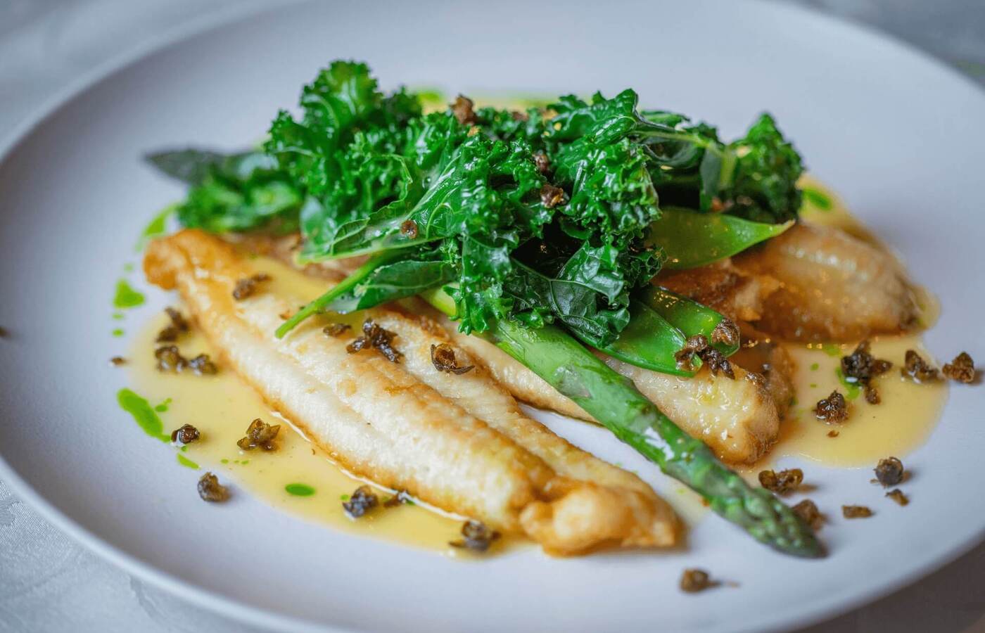 Smażone filety białej ryby ze szparagami, duszonym jarmużem, maślano‑cytrynowym sosem i chrupiącymi kaparami na białym talerzu