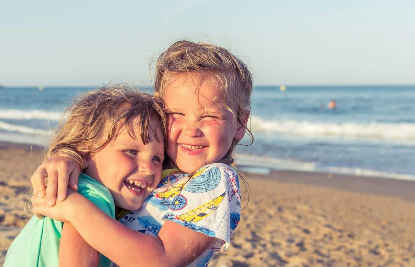 Dwie uśmiechnięte dzieci przytulają się na słonecznej piaszczystej plaży z łagodnymi falami, idealne dla rodzinnych wakacji.