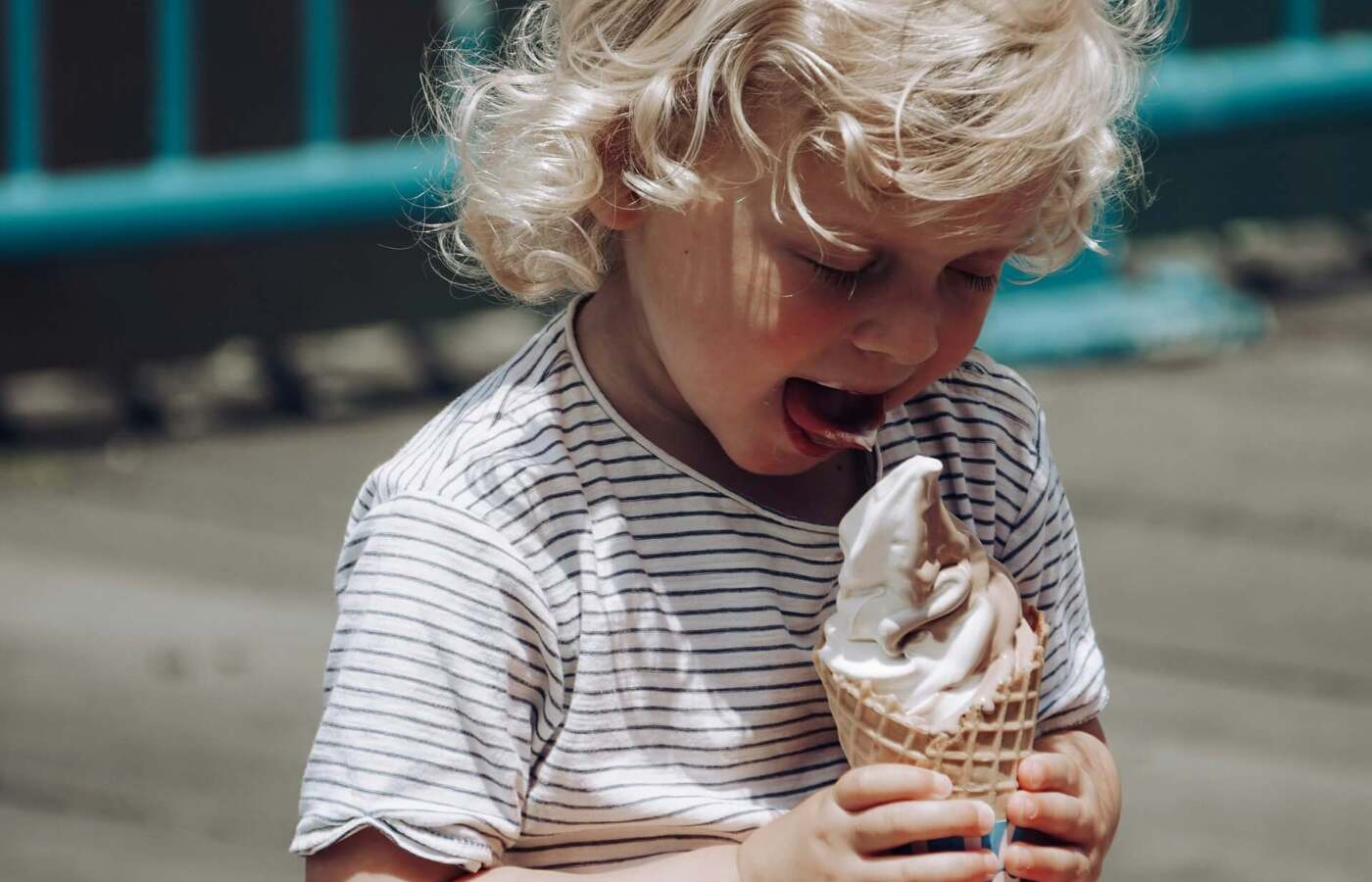 Dziecko o kręconych blond włosach siedzi na drewnianym deptaku, zajada czekoladowo‑waniliowe lody soft w wafelku