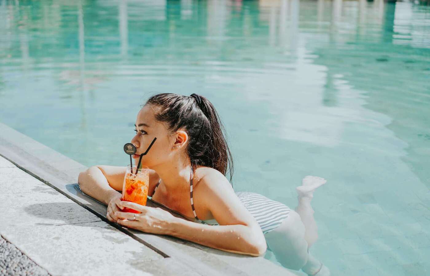 Kobieta relaksująca się na krawędzi turkusowego basenu hotelowego w pasiastym stroju, popijając jasny tropikalny koktajl