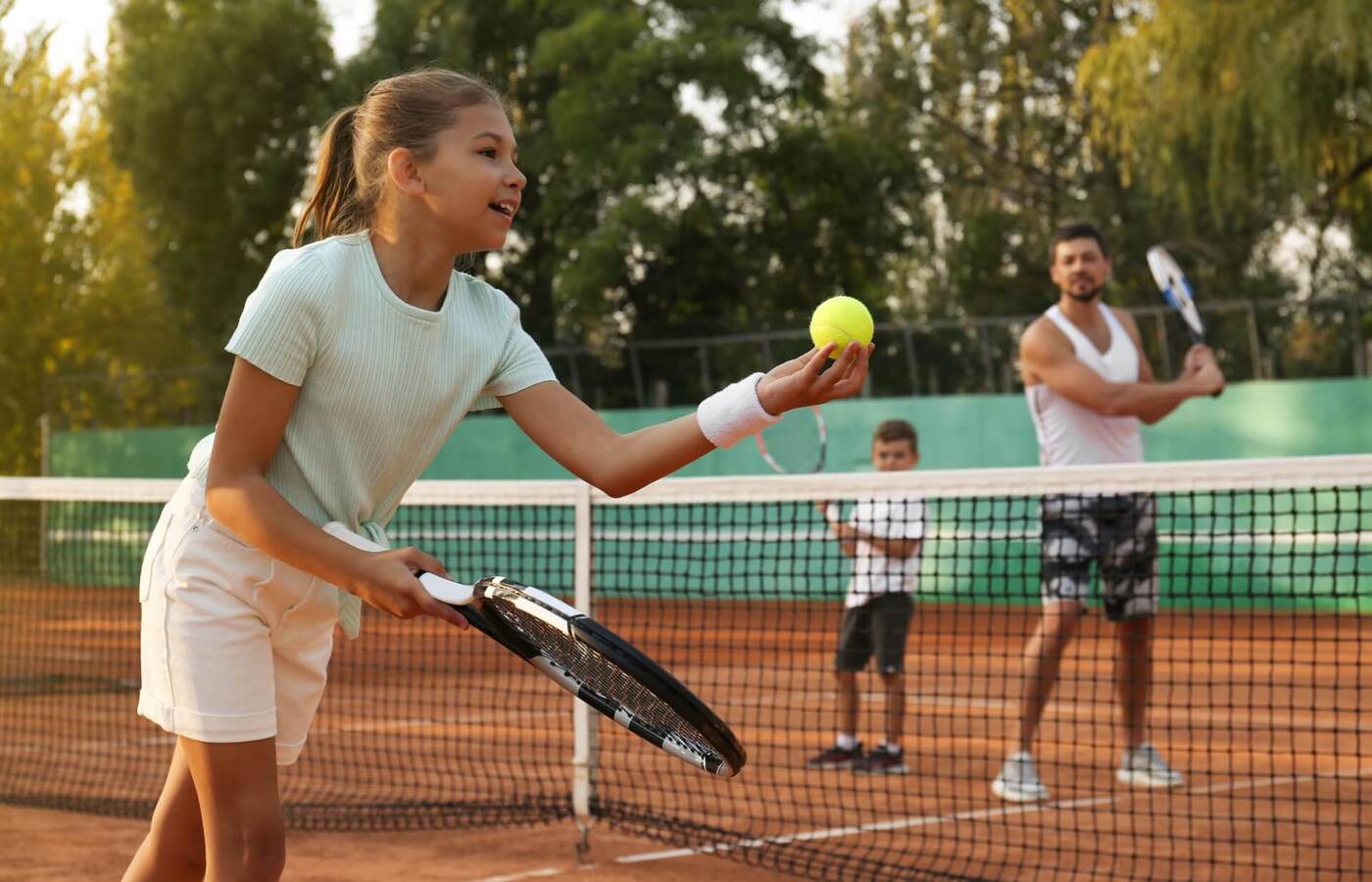 Rodzina gra na zewnętrznym korcie ziemnym — dziewczynka serwuje piłkę, ojciec i syn czekają za siatką, otoczenie drzew