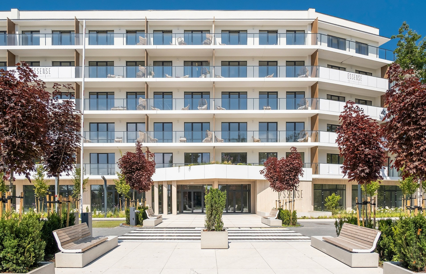 Nowoczesna fasada hotelu z przeszklonymi balkonami, wejściem do spa ESSENSE, zadbanym dziedzińcem i miejscami do siedzenia.