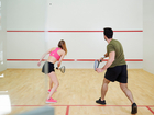 Two guests playing squash on bright hotel court with wooden floor, glass wall and red lines.