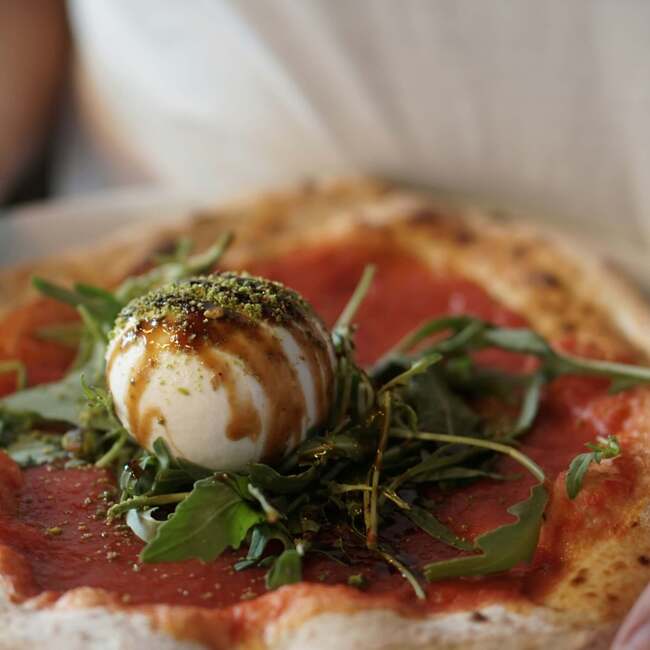 Rustykalna pizza z sosem pomidorowym, świeżą burratą, rukolą, glazurą balsamiczną i kruszonymi pistacjami na białym talerzu