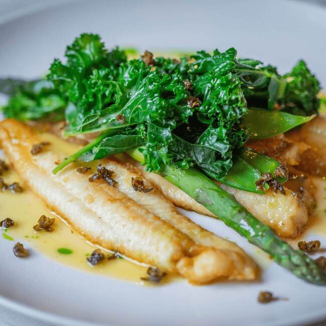 Fried white fish fillets with asparagus, stewed kale, buttery lemon sauce and crispy capers on white plate.