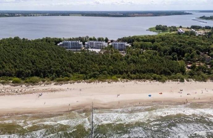 Niskie budynki nadmorskiego ośrodka za sosnowym lasem, szeroka piaszczysta plaża z parasolami, łagodne fale i falochron.