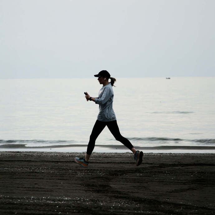 Osoba biegnąca w czapce i słuchawkach wzdłuż pustej plaży, spokojne fale i pochmurne niebo