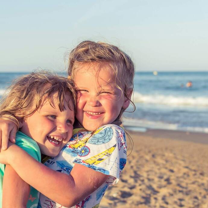 Dwie uśmiechnięte dzieci przytulają się na słonecznej piaszczystej plaży z łagodnymi falami, idealne dla rodzinnych wakacji.