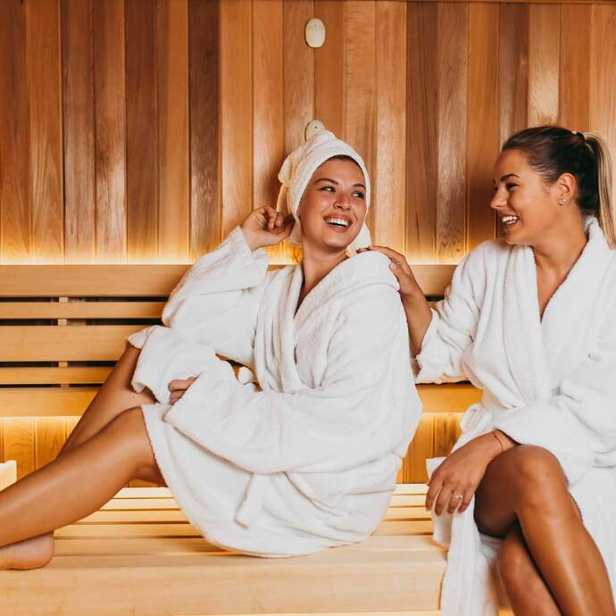 Dwie kobiety w białych szlafrokach relaksują się na drewnianych ławach sauny przy ciepłym, nastrojowym oświetleniu