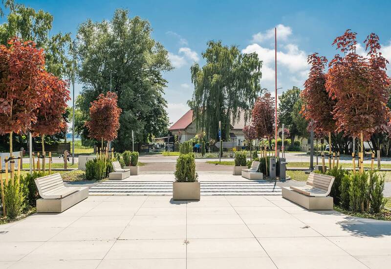 Słoneczne patio hotelowe z nowoczesnymi kamiennymi ławkami, donicami i czerwonymi drzewkami przy wyłożonej alejce prowadzącej do niskiego budynku.