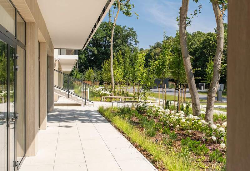 Nowoczesny taras hotelowy na parterze z przeszklonymi drzwiami, zacienionym przejściem i rabatami z białymi kwiatami i młodymi drzewami