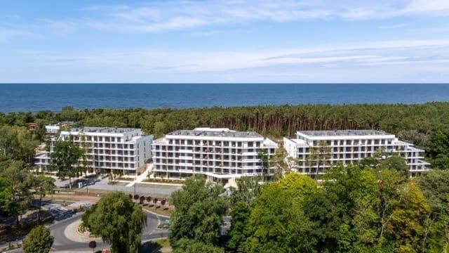 Nowoczesny nadmorski hotel z trzema białymi budynkami, balkonami, tarasami, odkrytym basenem, lasem sosnowym i widokiem na morze.