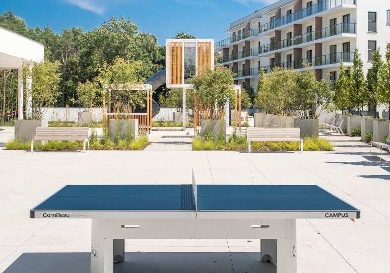 Stół do ping‑ponga na słonecznym dziedzińcu hotelu, nowoczesny budynek z balkonami, ławki i zadbana zieleń.