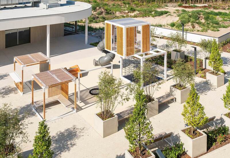 Nowoczesne patio hotelowe z zadaszonymi pergolami, modułowym placem zabaw z wieżą i zjeżdżalnią, ławkami i dużymi donicami
