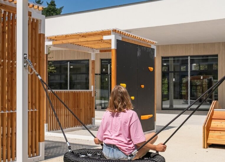 Młody gość na okrągłej huśtawce linowej na słonecznym patio hotelowym, pergole, ścianka wspinaczkowa i miejsca do siedzenia