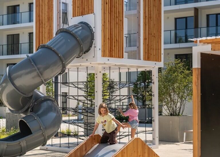 Dzieci bawią się na nowoczesnym, drewniano-metalowym placu zabaw z krętą, zamkniętą zjeżdżalnią i linową ścianką na hotelowym, słonecznym dziedzińcu