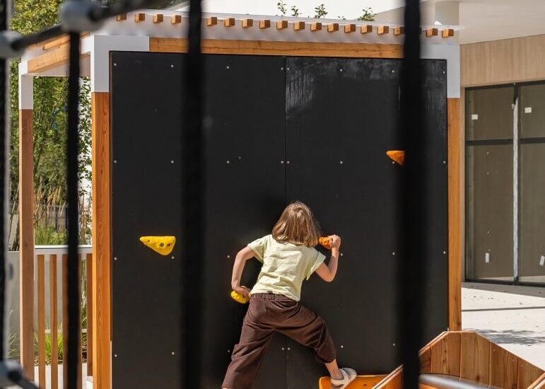 Dziecko wspinające się na czarną ściankę z żółtymi chwytami w słonecznym, drewnianym placu zabaw hotelu