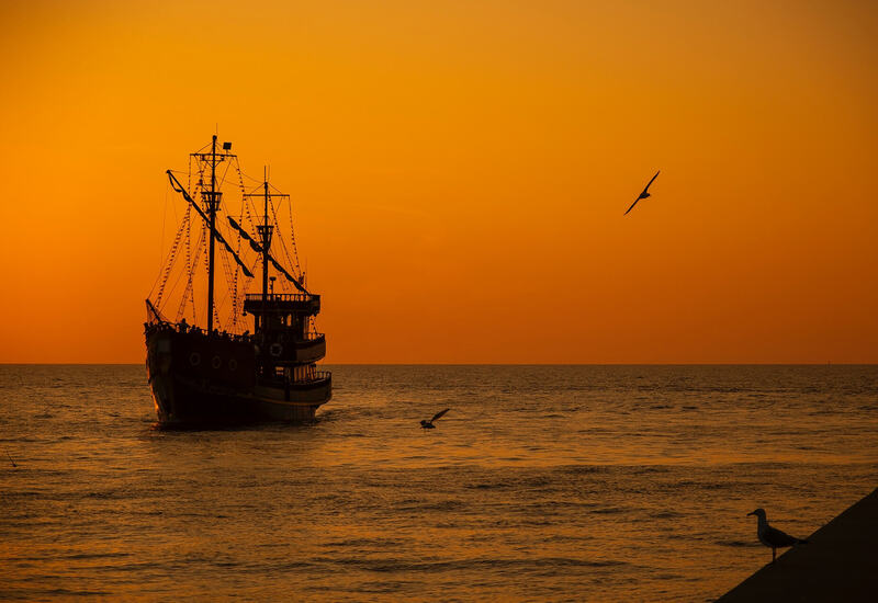 Sylwetka statku wycieczkowego na spokojnym morzu podczas pomarańczowego zachodu słońca, mewy w locie i na molo, romantyczny wieczorny rejs