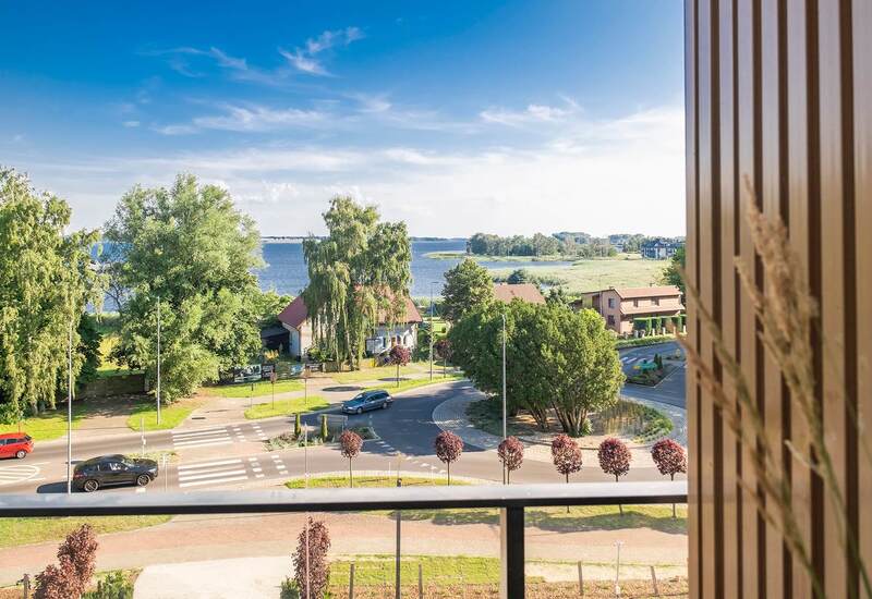 Widok z balkonu na zadrzewioną ulicę i rondo, z panoramą spokojnego jeziora, domów i zielonego brzegu