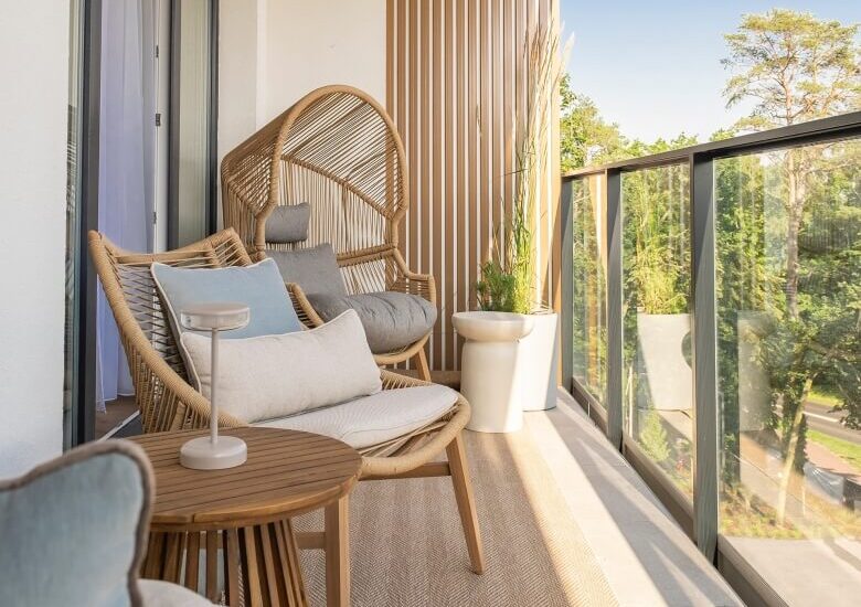 Prywatny balkon z rattanowymi fotelami, miękkimi poduszkami, małym stolikiem i szklaną balustradą oraz widokiem na drzewa i słońce.
