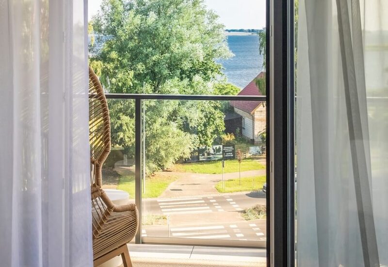 Prywatny balkon z wiklinowym fotelem, przeszklonym balustradą i lekkimi zasłonami, z widokiem na zieleń i wodę.