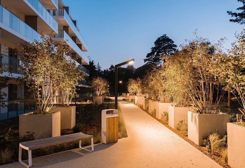 Zaciszny dziedziniec hotelu o zmierzchu z oświetloną ścieżką, dużymi donicami, ławkami i szklanymi balkonami pokoi