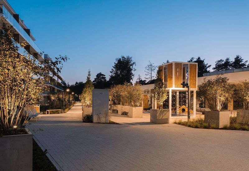 Nowoczesne patio hotelowe o zmierzchu z oświetlonymi drzewami, brukowanym deptakiem, ławkami i drewnianą altaną przy leśnym tle.