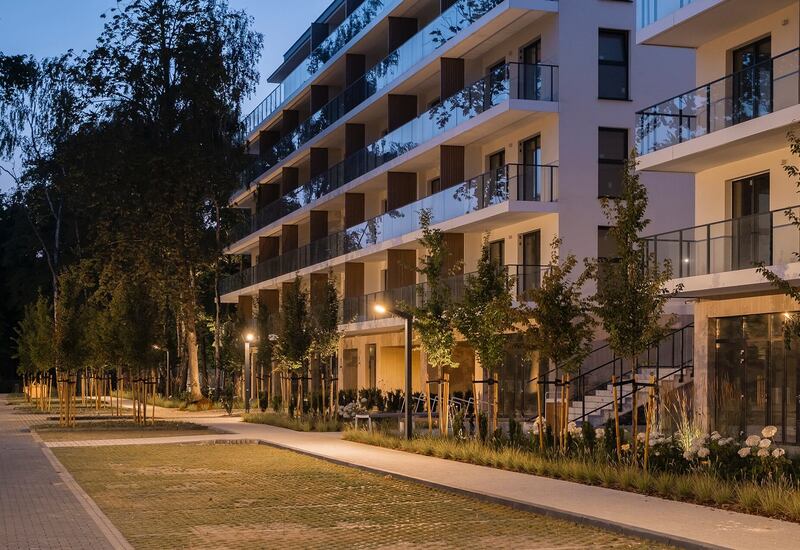 Nowoczesny hotel z balkonami i szklanymi balustradami, oświetlony drzewami deptak wieczorem, tarasy na parterze i zadbana zieleń.