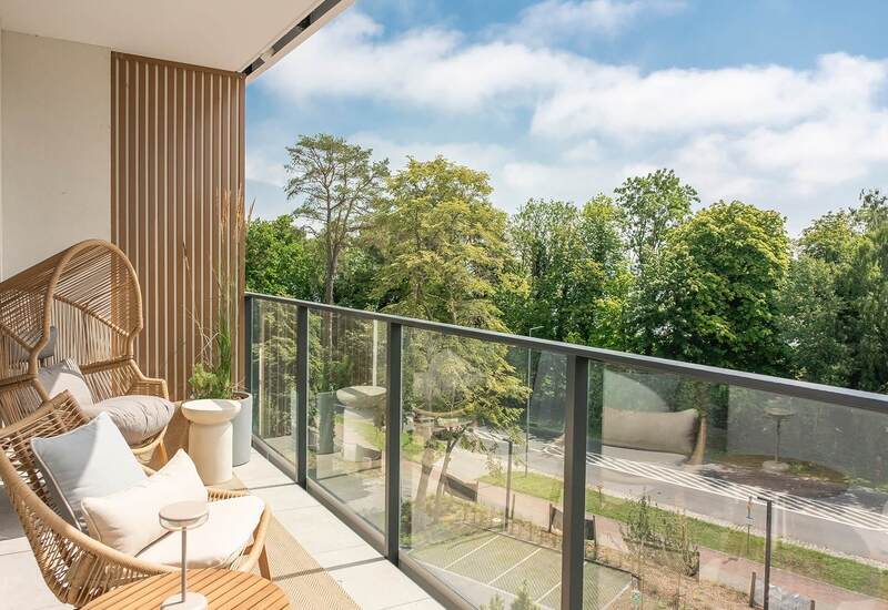 Prywatny balkon z rattanowymi fotelami, poduszkami i stolikiem, ze szklaną balustradą i widokiem na zielone korony drzew