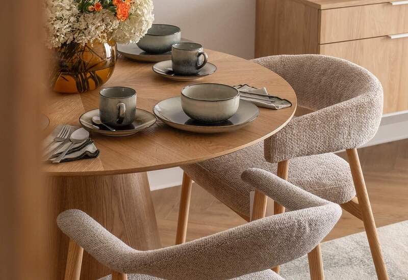 Okrągły dębowy stół z zastawą dla czterech, miękkie tapicerowane krzesła i bukiet świeżych kwiatów.