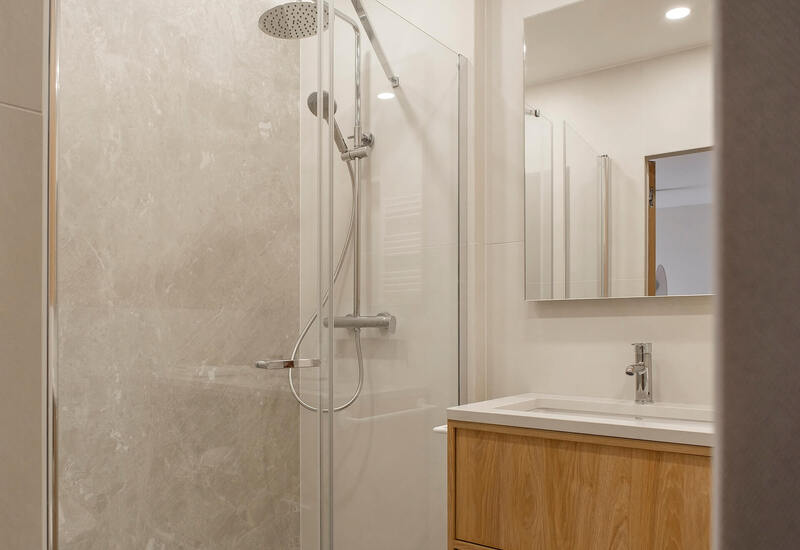 Nowoczesna łazienka hotelowa z przeszkloną kabiną prysznicową, deszczownicą i słuchawką, marmurowymi płytkami oraz drewnianą szafką z umywalką.