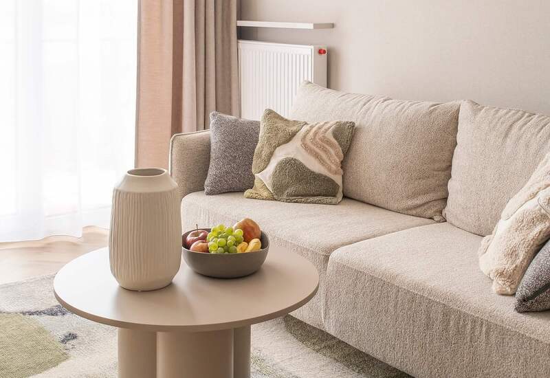 Strefa wypoczynkowa w apartamencie: beżowa sofa z dekoracyjnymi poduszkami, okrągły stolik z wazonem i miską owoców, lekkie zasłony i ciepłe lampy.