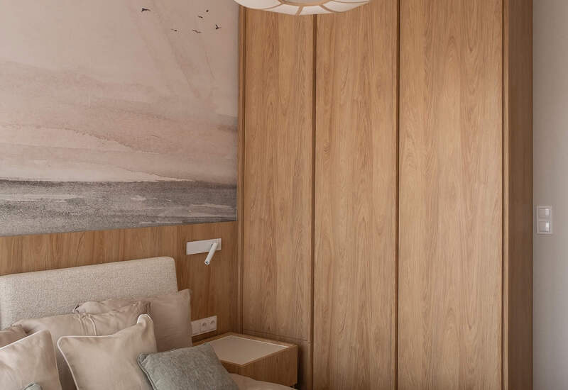 Przytulny pokój z neutralną pościelą, miękkim zagłówkiem, stolikiem nocnym z lampką, drewnianą szafą i wiszącą lampą.