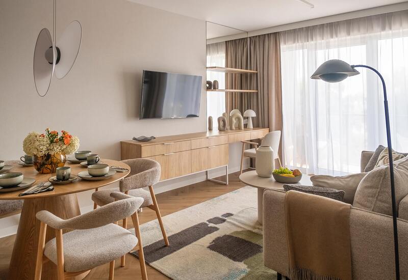 Jasny, nowoczesny salon w apartamencie z nakrytym stołem i wazonem kwiatów, TV na ścianie, drewniana komoda, firany i przytulna sofa.