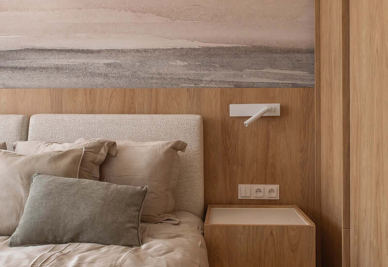 Sypialnia w neutralnych tonach z tapicerowanym zagłówkiem, drewnianą szafką nocną, kinkietem do czytania, miękką pościelą i muralem morza