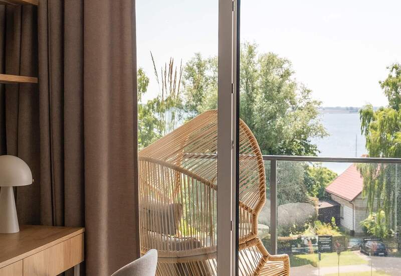 Prywatny balkon z plecionym fotelem rattanowym i szklaną balustradą, widok na jezioro i drzewa; wnętrze z biurkiem, lampą i wygodnym krzesłem.