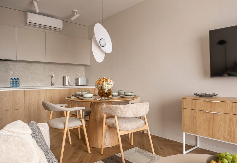 Jadalniany kącik w apartamencie: okrągły drewniany stół dla czterech, wisząca lampa, aneks kuchenny, klimatyzacja i telewizor.