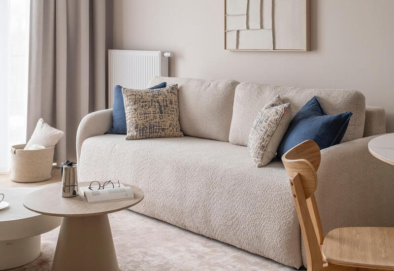 Kącik w apartamencie hotelowym z kremową sofą boucle, wzorzystymi i granatowymi poduszkami, okrągłymi stolikami, ekspresem, dywanem i zasłonami