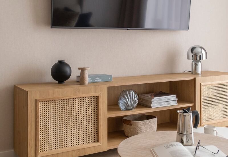 Strefa wypoczynkowa z TV na ścianie, drewniana szafka z rattanowymi frontami, okrągły stolik z książką i kawiarką.