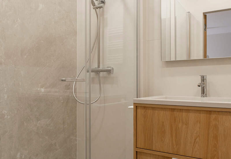 Nowoczesna łazienka hotelowa z przeszklonym prysznicem, deszczownicą i słuchawką, marmurową ścianą i drewnianą szafką z umywalką.