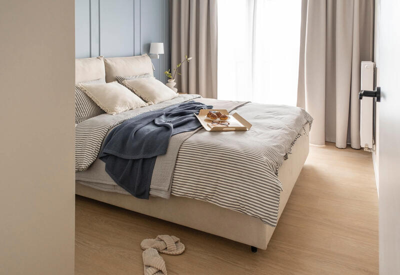 Jasny, stonowany pokój hotelowy z dużym łóżkiem, pasiastą pościelą i granatowym pledem, tacą ze śniadaniem, lampką i kapciami na drewnianej podłodze