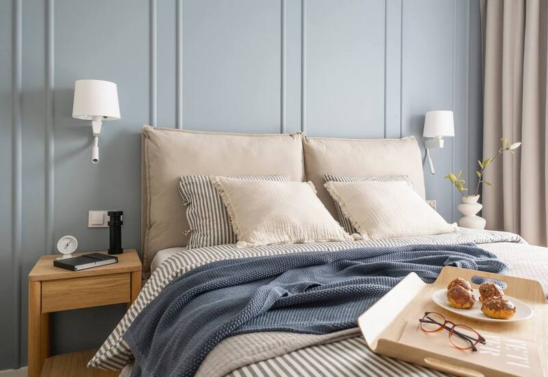 Łóżko dwuosobowe z tapicerowanym zagłówkiem, niebieska boazeria, pasiasta pościel i dziany koc, lampki, taca z ciastkami i okularami