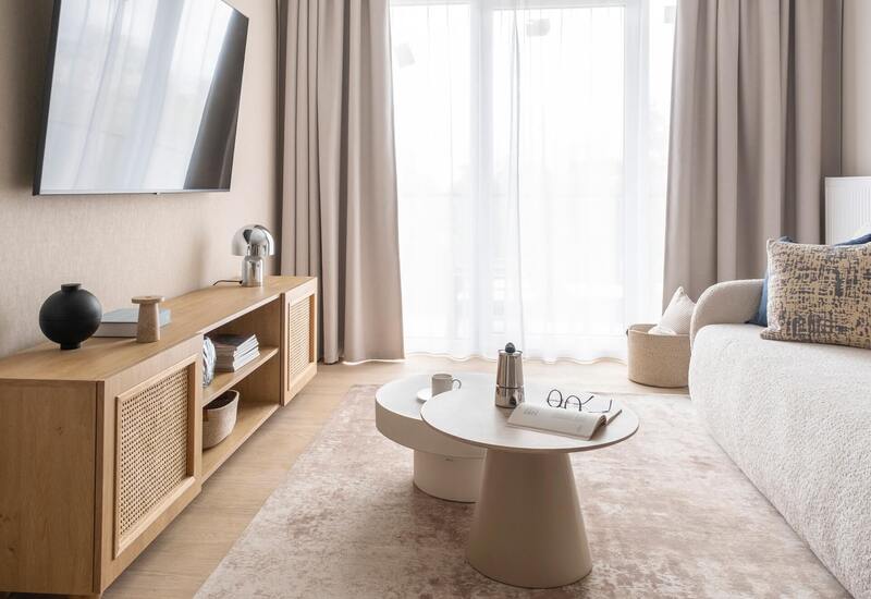 Jasny salon apartamentu z miękką sofą, poduszkami, telewizorem na ścianie, drewnianą szafką, okrągłymi stolikami i kawiarką.