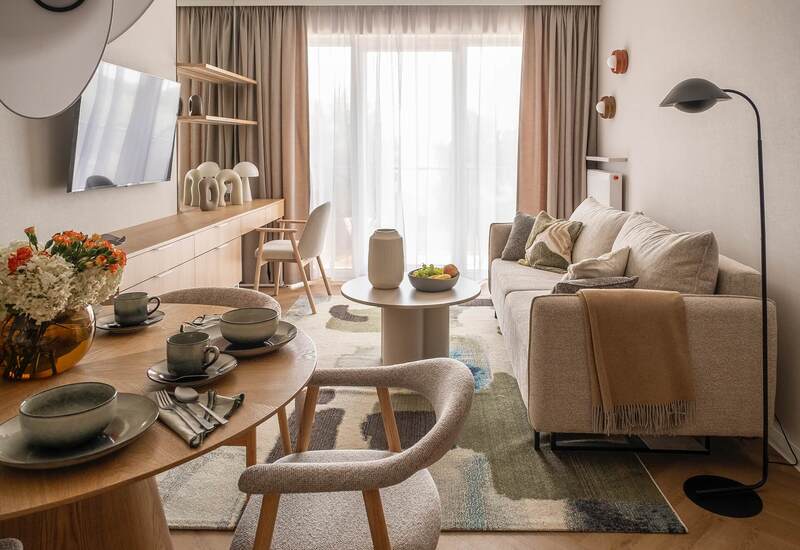 Nowoczesny apartament: beżowa sofa, okrągły stolik z owocami, jadalnia dla dwóch, biurko, telewizor i światło z balkonu.