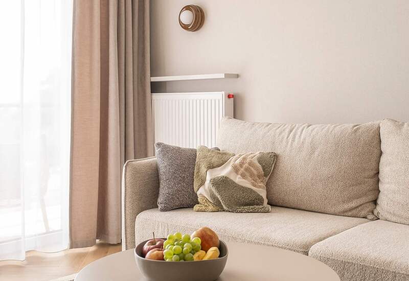 Jasny salon hotelowy z beżową sofą i dekoracyjnymi poduszkami, okrągły stolik z miską owoców, przezroczyste zasłony i kinkiety