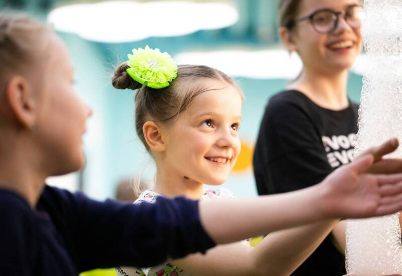 Uśmiechnięte dzieci w klubie dla rodzin bawią się baniek; dziewczynka z zieloną kwiatową spinką we włosach.