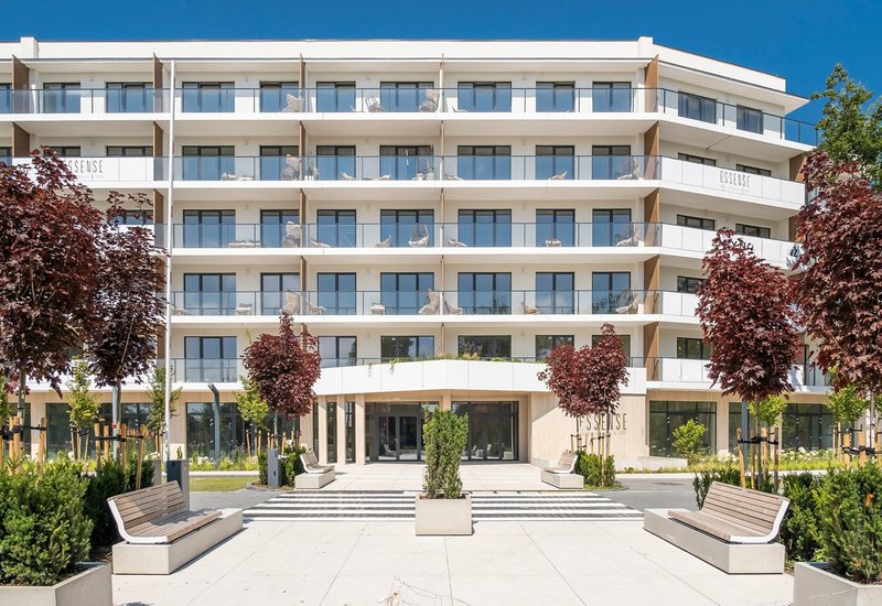 Nowoczesna fasada hotelu z przeszklonymi balkonami, wejściem do spa ESSENSE, zadbanym dziedzińcem i miejscami do siedzenia.