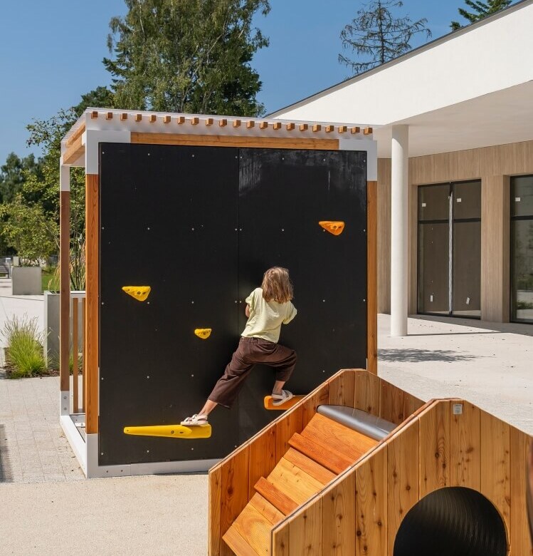 Zewnętrzny plac zabaw dla rodzin, dziecko na czarnej ściance wspinaczkowej z żółtymi chwytami i drewnianym tunelem przy nowoczesnym hotelu