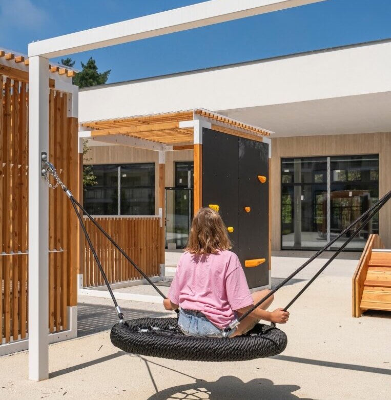 Młody gość na okrągłej huśtawce linowej na słonecznym patio hotelowym, pergole, ścianka wspinaczkowa i miejsca do siedzenia