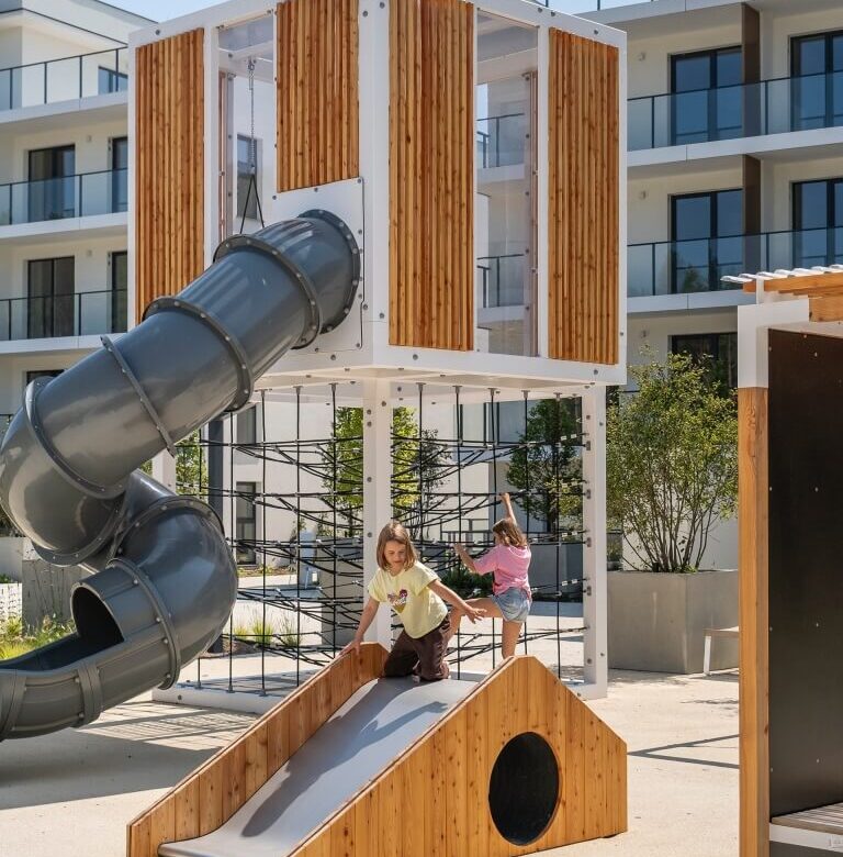 Dzieci bawią się na nowoczesnym, drewniano-metalowym placu zabaw z krętą, zamkniętą zjeżdżalnią i linową ścianką na hotelowym, słonecznym dziedzińcu
